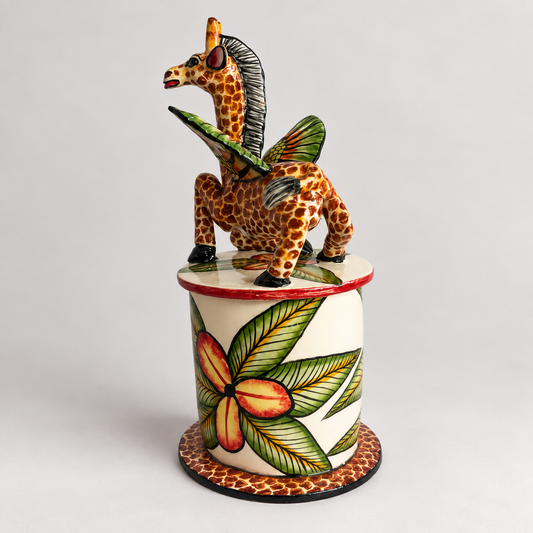 A hand-painted ceramic figurine of a giraffe with green wings, perched on top of a cylindrical pedestal decorated with a large green and orange flower, set against a plain light gray background.