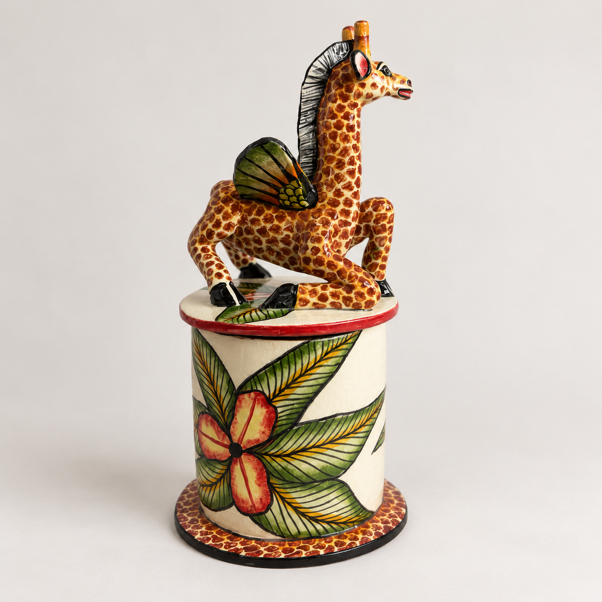 A hand-painted ceramic figurine of a giraffe with wings, perched on top of a cylindrical pedestal decorated with green leaves and red flowers, set against a plain light gray background.
