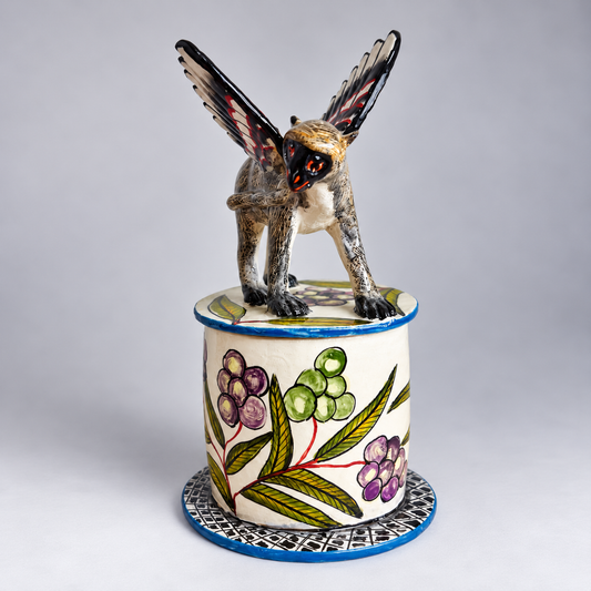 A hand-painted ceramic sculpture of a winged monkey atop a cylindrical pedestal decorated with botanical motifs, set against a neutral gray studio background.