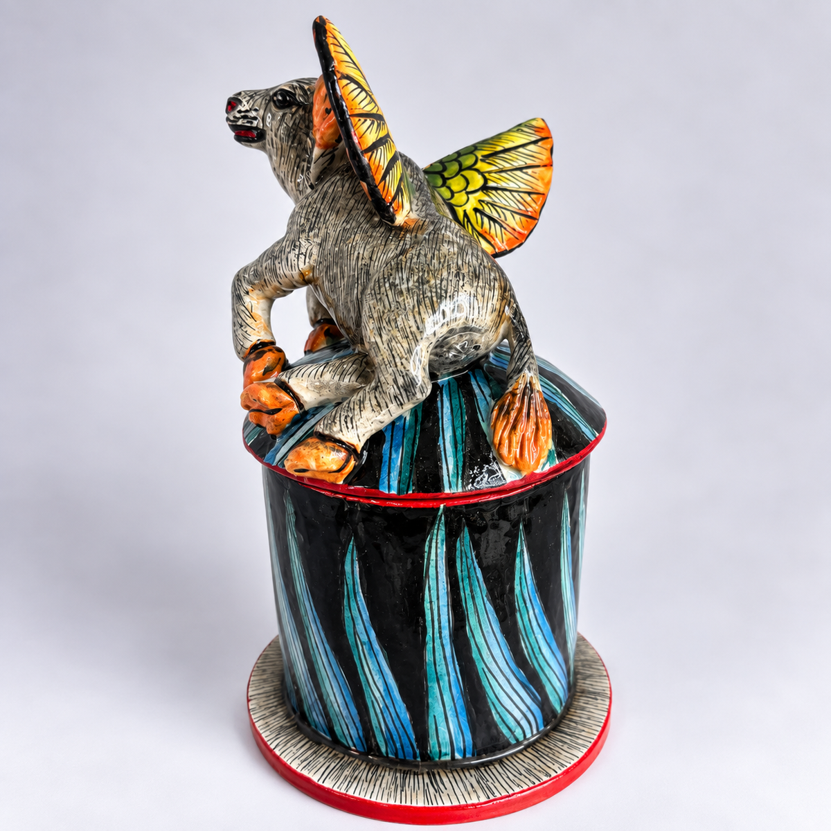 A hand-painted ceramic figurine of a fantasy buffalo with orange wings and hooves, perched atop a tall black and blue striped pot with red trim, set against a plain light gray background.