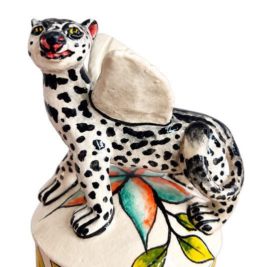 A ceramic trinket jar shaped. Lid has a spotted big cat, shown standing on a floral base against a light gray background.