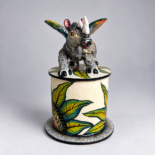 A hand-painted ceramic lidded box featuring a detailed rhinoceros figurine on top. The cylindrical box is decorated with green and yellow tropical leaves on an off-white background, and the lid is topped with the rhinoceros sculpture showing black, white, and gray textures with colorful accents.