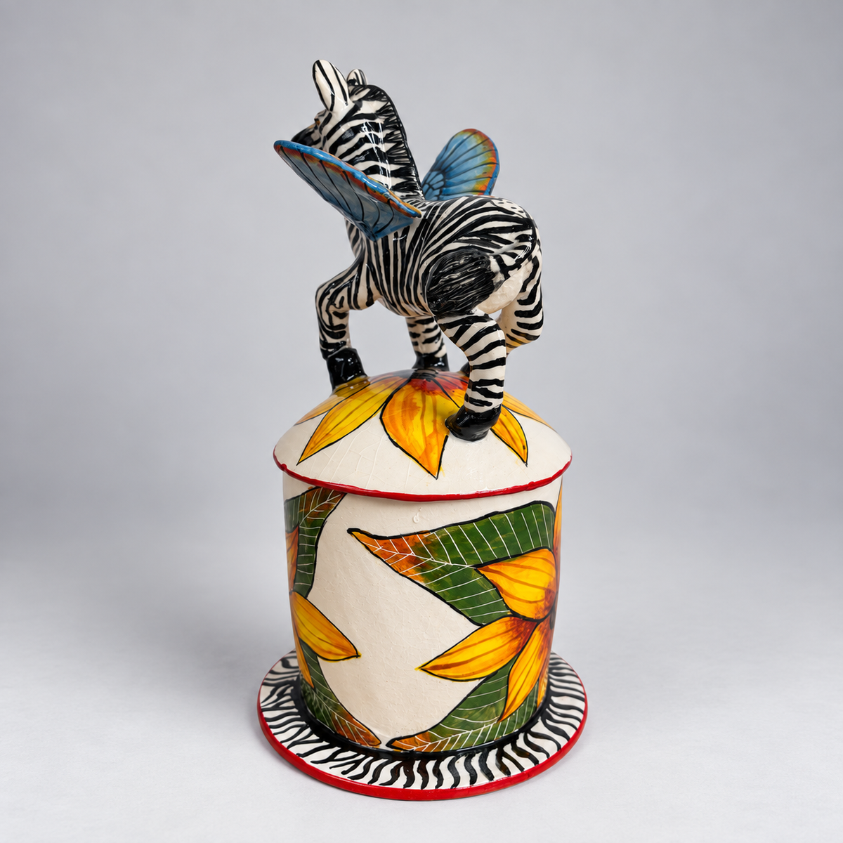 A handcrafted ceramic sculpture of a zebra standing on top of a cylindrical base decorated with yellow and green tropical leaves. The zebra has black and white stripes with blue wings, set against a neutral gray background.