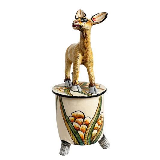 A hand-painted ceramic figurine of a small brown deer standing on top of a cylindrical cream-colored pot decorated with yellow, green, and orange floral patterns, set against a plain light gray background.