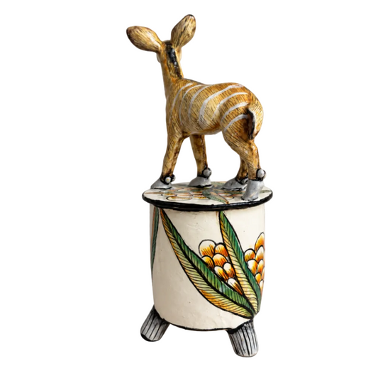 A hand-painted ceramic figurine of a zebra standing on top of a decorated cylindrical pedestal, photographed against a neutral light gray background.