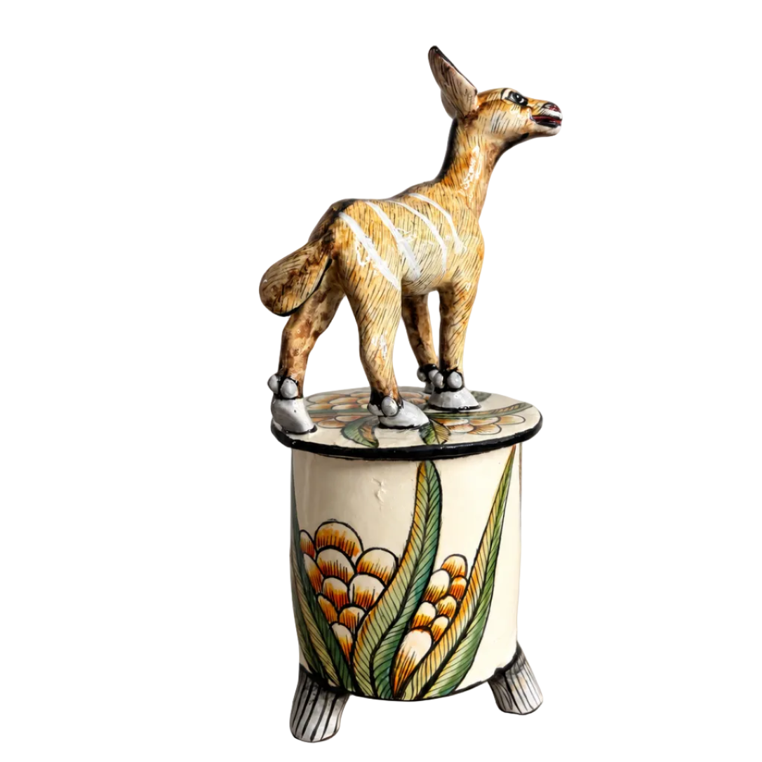A decorative ceramic figurine of a deer standing on top of a cylindrical pedestal with floral patterns, photographed against a neutral gray studio background.