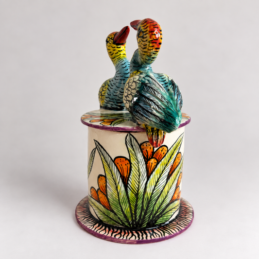 A colorful ceramic bird figurine perched on top of a cylindrical floral pot. The birds are painted with yellow, green, blue, and red patterns, while the pot features green leaves and orange flowers on a cream-colored base, all set against a plain light gray background.
