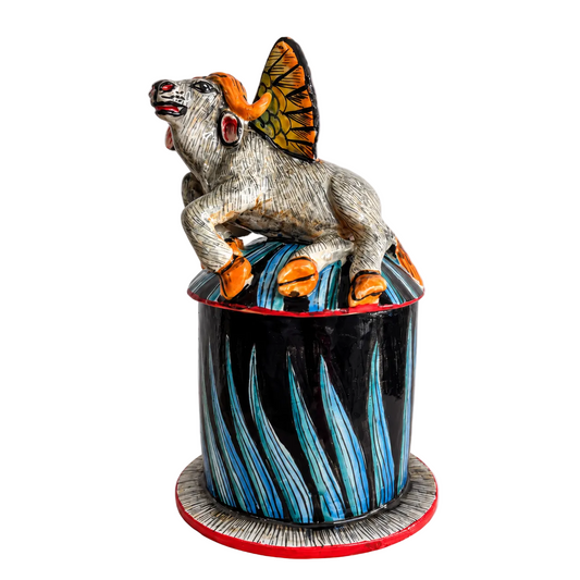 A hand-painted ceramic figurine of a buffalo perched atop a tall cylindrical pot. The buffalo has a textured gray body, bright orange hooves and horns, and a patterned orange and yellow wings.