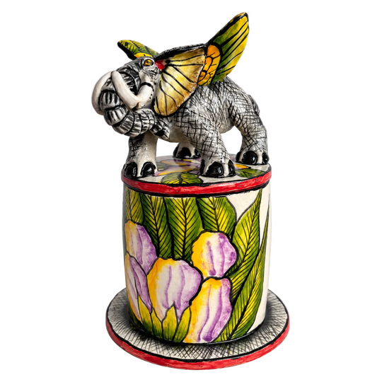 A hand-painted ceramic sculpture of an elephant with colorful wings standing on top of a cylindrical pot decorated with purple and yellow flowers and green leaves.