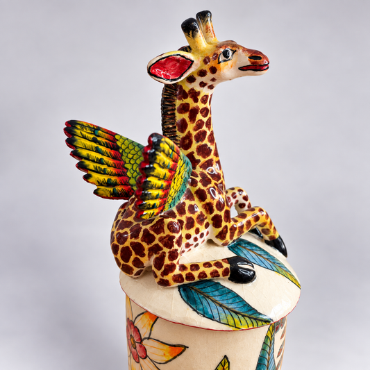 A hand-painted ceramic giraffe figurine with colorful wings, perched on top of a decorated cylindrical base against a neutral gray background.