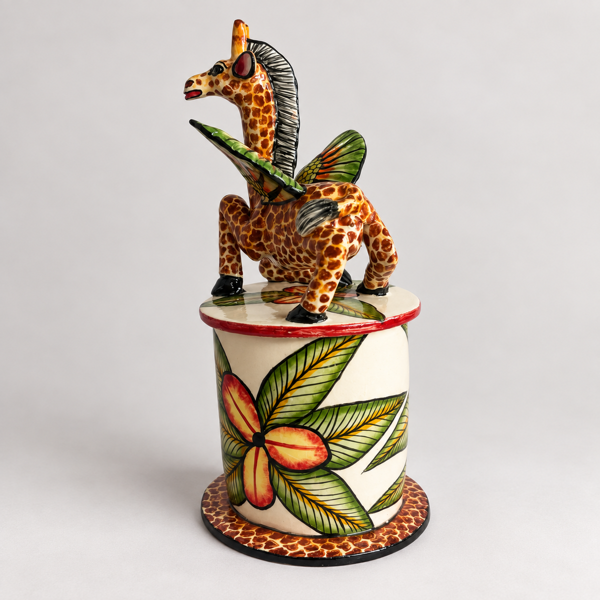 A hand-painted ceramic figurine of a giraffe with green wings, perched on top of a cylindrical pedestal decorated with a large green and orange flower, set against a plain light gray background.