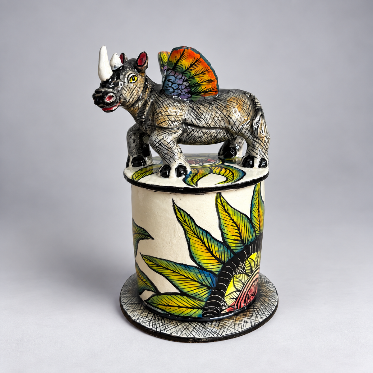 A handcrafted ceramic sculpture of a rhinoceros with a colorful winged back, standing atop a cylindrical pot decorated with green tropical leaves, set against a neutral gray background.