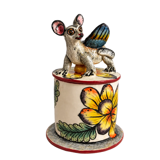 A handcrafted ceramic figurine of a winged creature perched atop a cylindrical lidded box decorated with a large yellow and orange flower and green leaves on a light gray background.