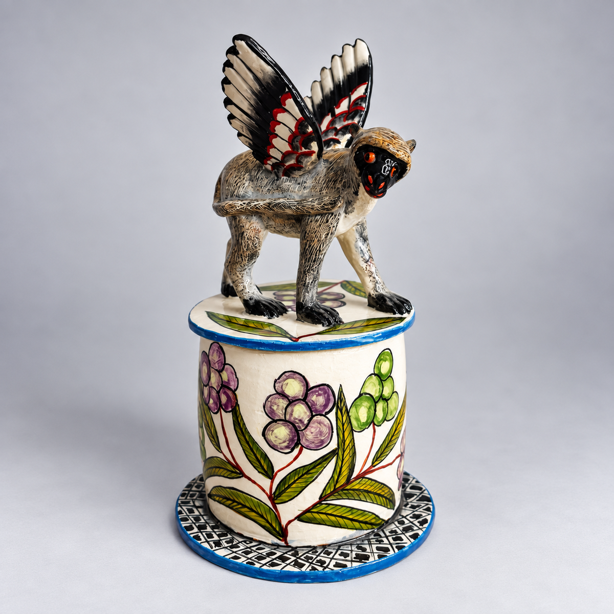 A hand-painted ceramic figurine of a winged monkey perched atop a decorated cylindrical pedestal, set against a neutral gray studio background.