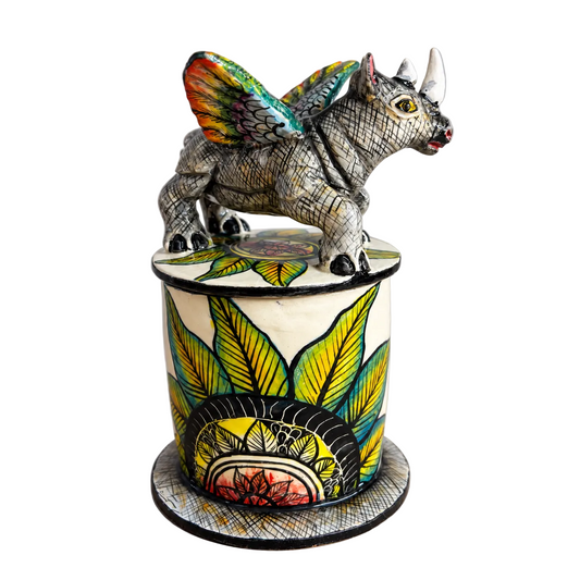 A handcrafted ceramic figurine of a winged rhinoceros standing atop a decorated cylindrical pedestal, photographed against a neutral light gray background.
