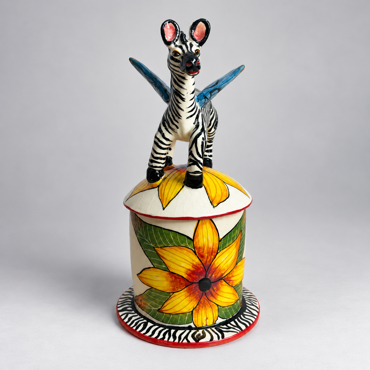 A hand-painted ceramic figurine of a zebra with blue wings standing on top of a floral pot. The zebra has black and white stripes with pink inner ears, and the pot features a large yellow flower with green leaves on a white background, all set against a neutral gray studio backdrop.