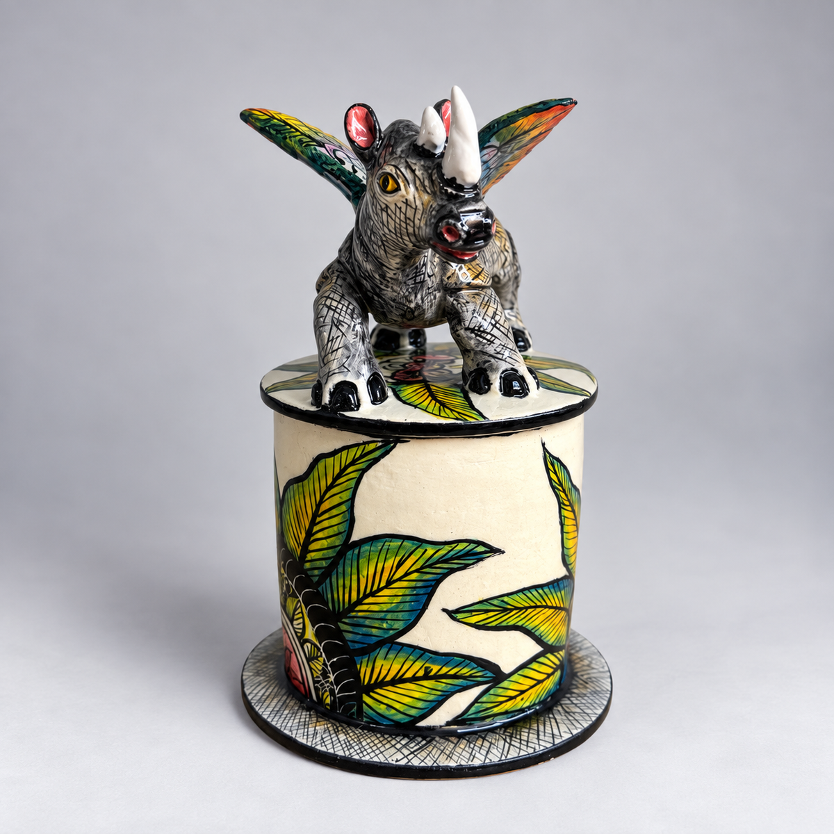 A hand-painted ceramic lidded box featuring a detailed rhinoceros figurine on top. The cylindrical box is decorated with green and yellow tropical leaves on an off-white background, and the lid is topped with the rhinoceros sculpture showing black, white, and gray textures with colorful accents.