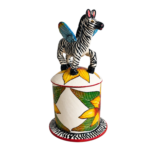 A hand-painted ceramic figurine of a Pegasus zebra with colorful wings, standing on top of a cylindrical lidded box decorated with yellow flowers and green leaves, set against a neutral gray background.