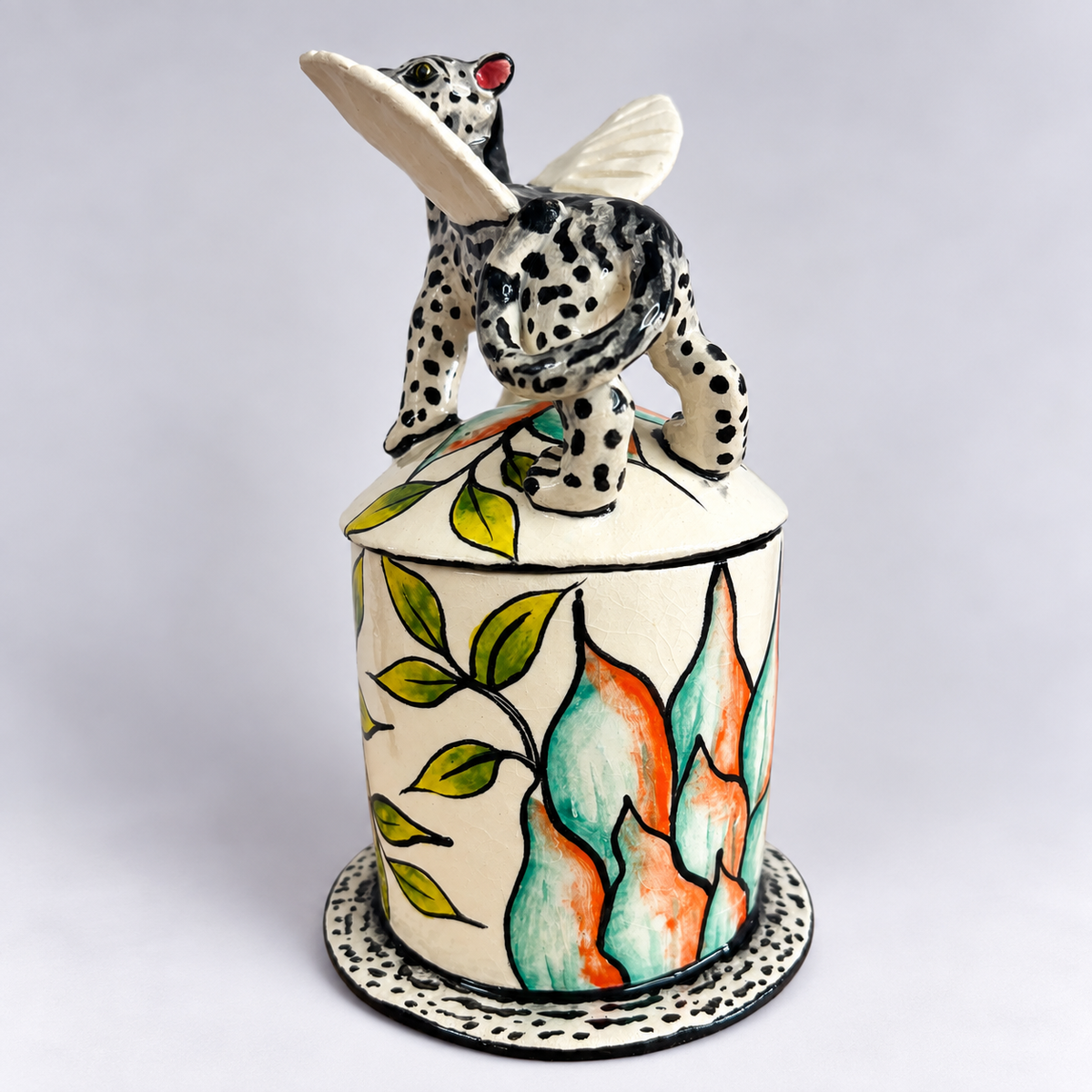 A hand-painted ceramic lidded jar featuring a white base decorated with green leaves and orange and teal flames. The lid is topped with a black and white spotted animal figurine with white wings.