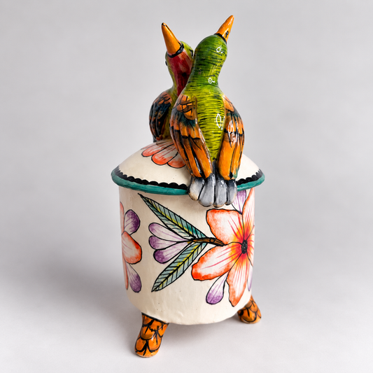 A hand-painted ceramic lidded container with two small birds perched on top. The cylindrical body is cream-colored with a large orange hibiscus flower and green leaves, while the lid features two birds with green, orange, and brown feathers.